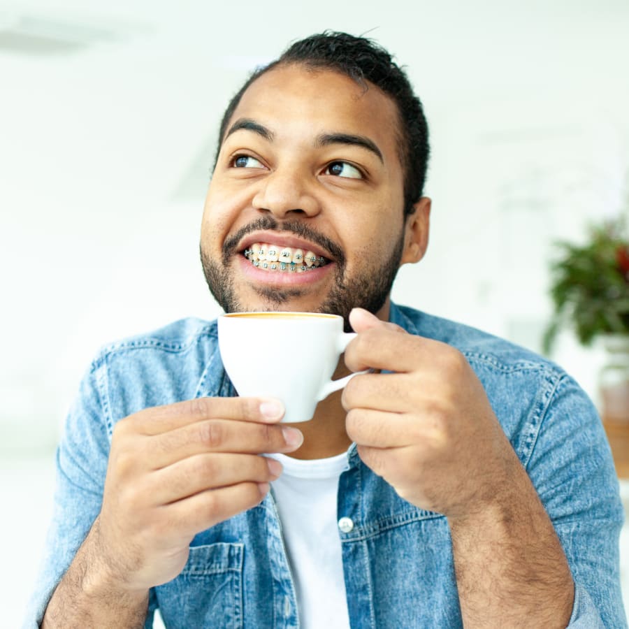 A man with braces wearing an open blue demin button up shirt with a white undershirt holds up a small white coffee cup. He is smiling and looking to his right.