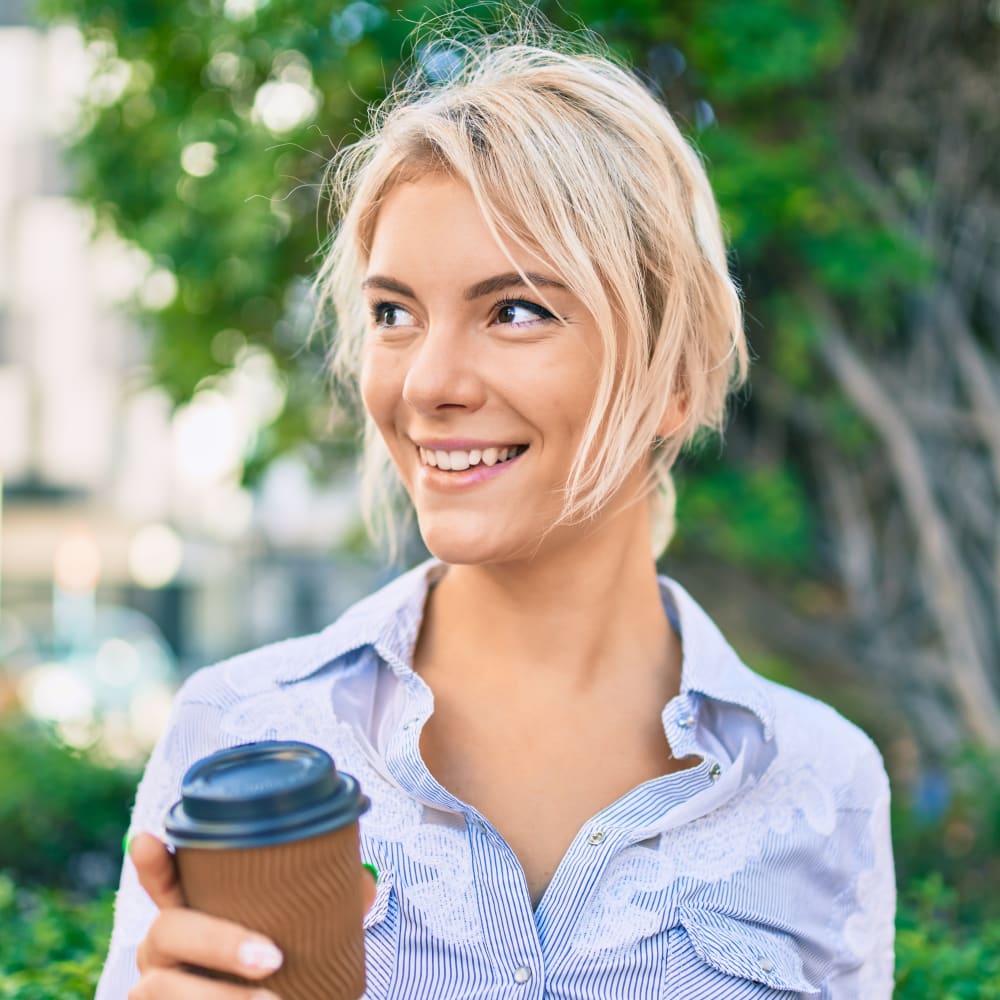 A woman wearing a blue and white thin striped shirt stands outside in front of some green trees while holding a brown cardboard coffee cup. She is smiling and looking to her right.