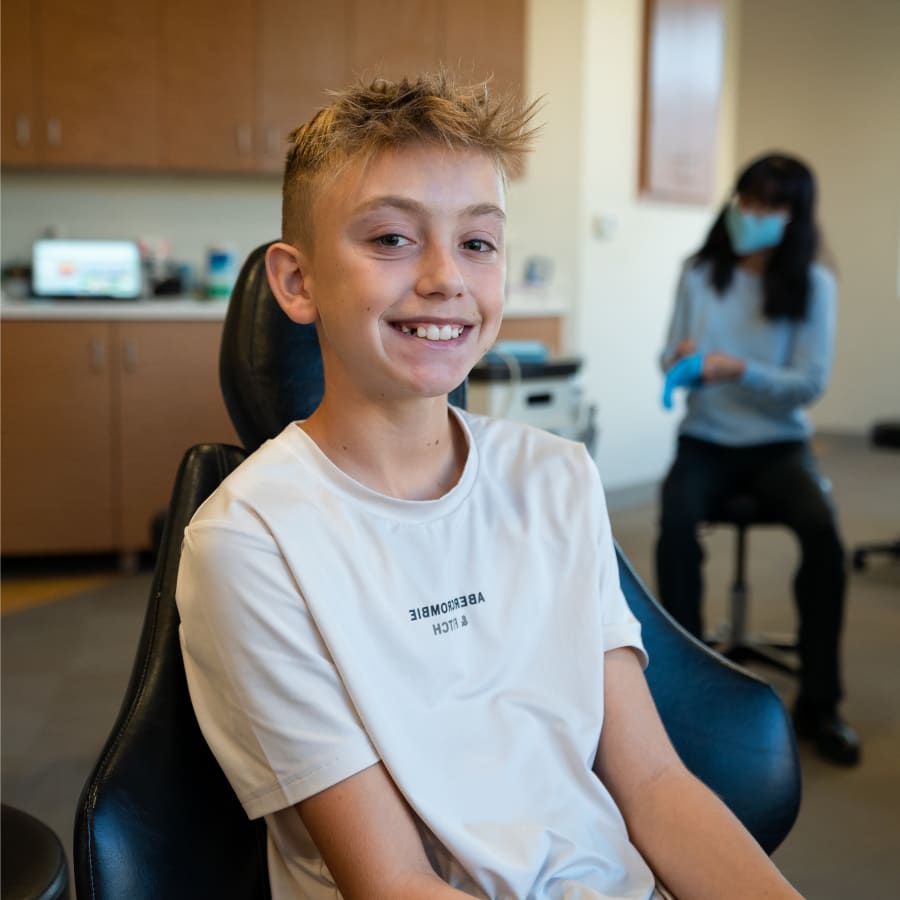 A young boy wearing a white t-shirt is sitting in a chair in a dentist's office smiling at the camera.