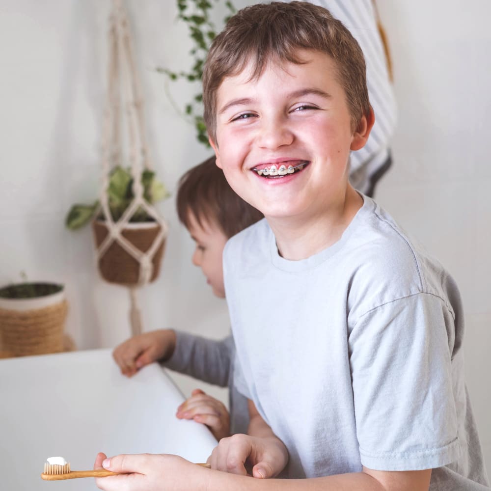 One boy stands in front of another boy at the bathroom sink. The boy in front has braces and is holding a toothbrush with toothpaste on it while smiling.