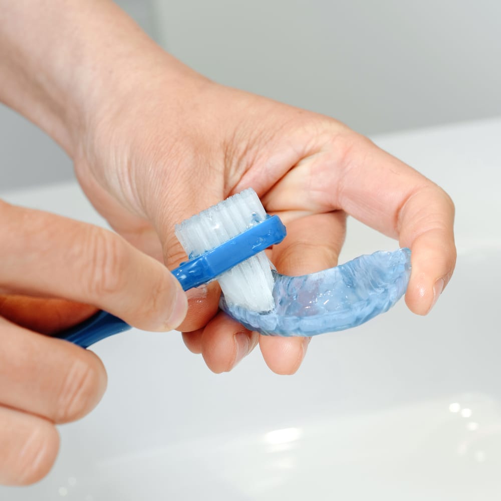 A woman and man sit at a table outside. The man's back is facing the camera with both of his hands on the table touching a coffee cup and the woman is looking at the man with her left hand up sitting under her chin.