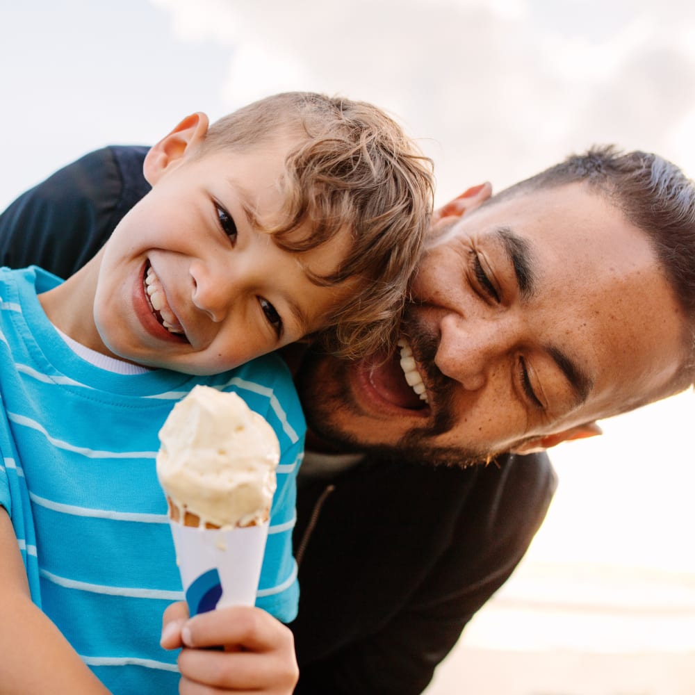 A boy wearing a bright blue shirt is holding a vanill ice cream cone while playfully avoiding a man who is standing behind and leaning over him trying to get to the ice cream.