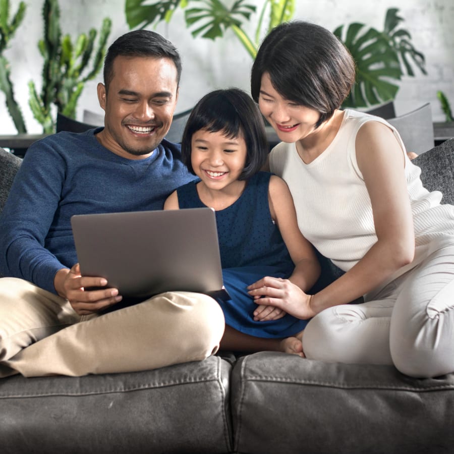 A family sitting outside with green trees in the background. The young boy is sitting on his fathers lap while the mother sits to the left of them with the sister to the left of the mother.