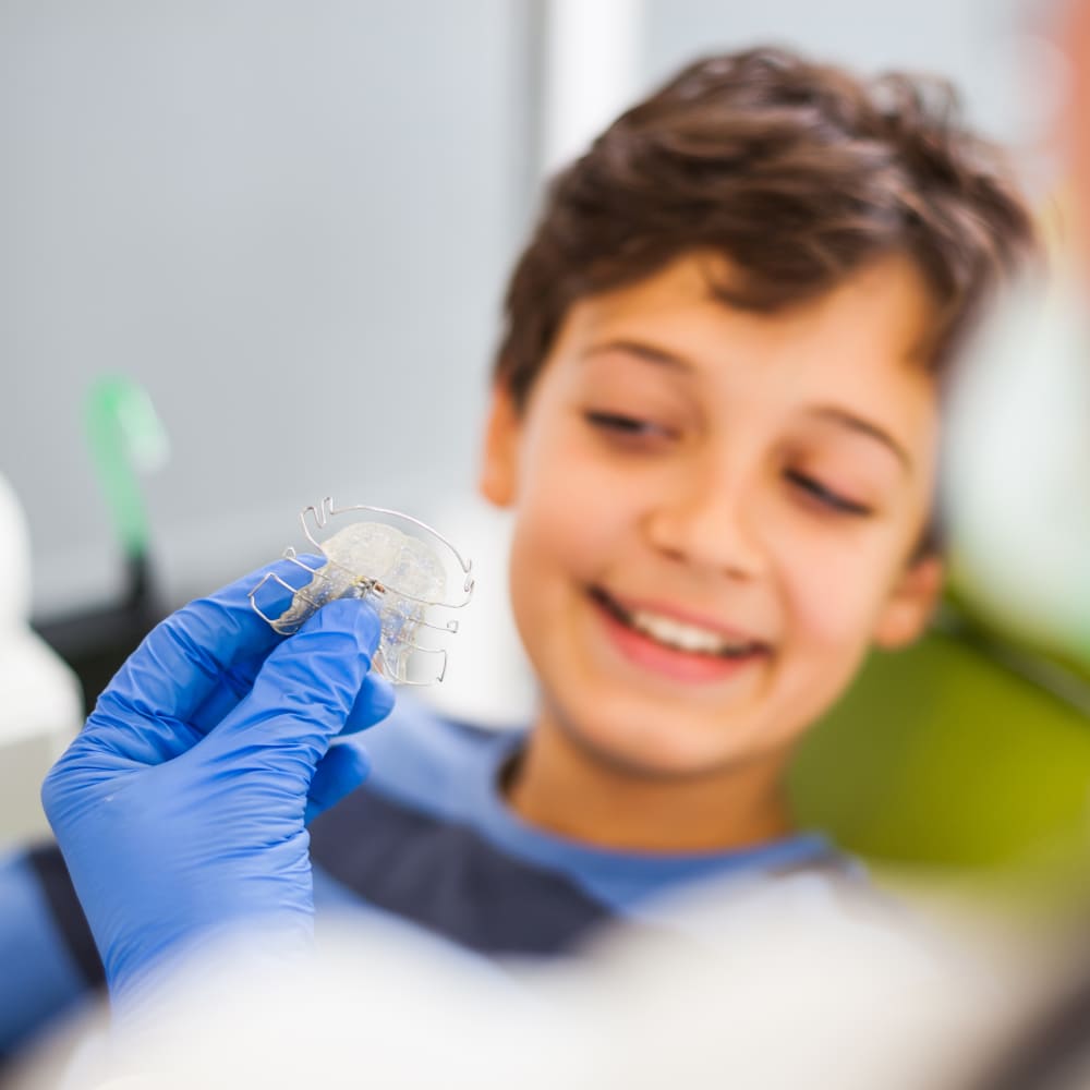A teenage girl with braces and a teenage boy sit on the floor holding video game controllers hile another teenage boy and girl sit on the couch just behind them also holding video game controllers.