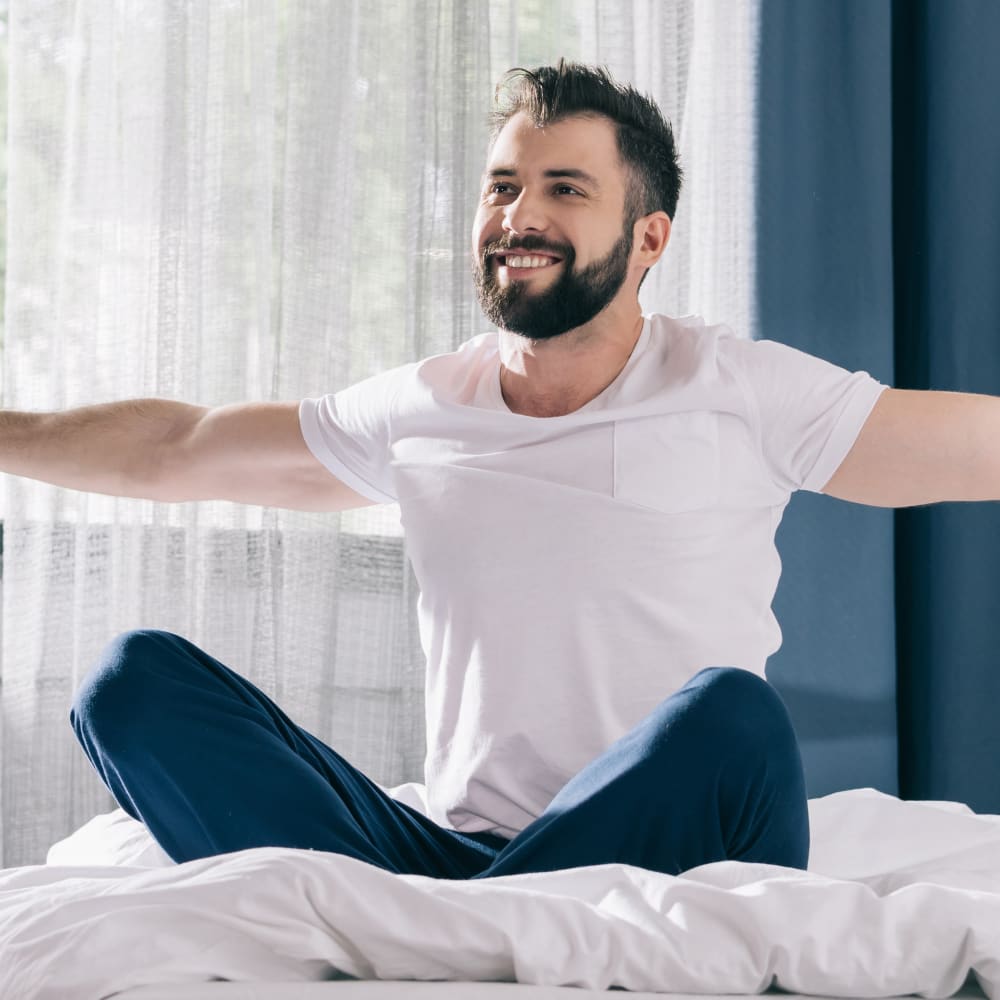 A man wearing a hite shirt and blue pants witting cross-legged on a bed with white blankets stretching his arms out on either side. There is a window in the background with white sheer curtains and a dark blue wall.