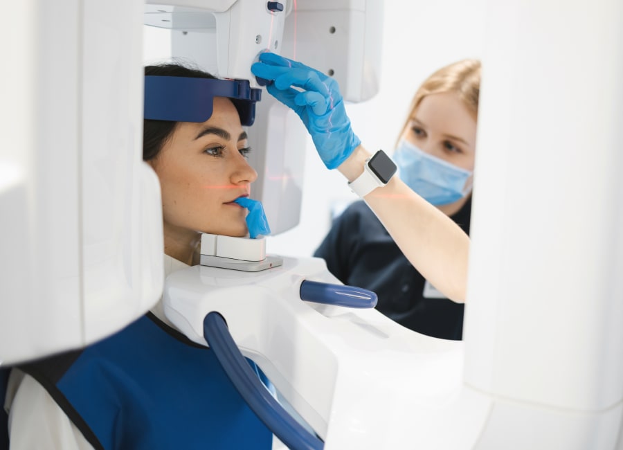 Dental technician setting up a 3D X-ray machine while a patient wearing a blue protective vest stands in the machine.