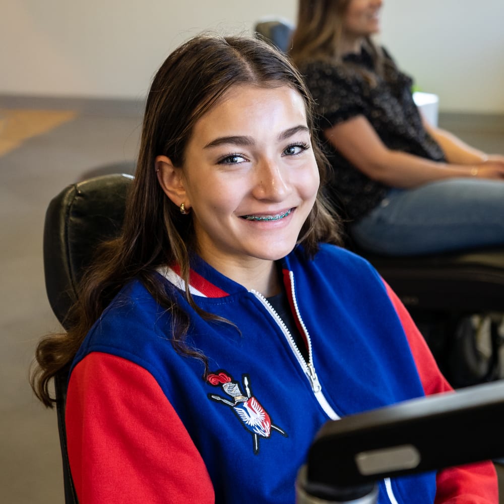 A teenage girl sits in a dentist's chair and smiles at the camera, displaying a full set of traditional braces.