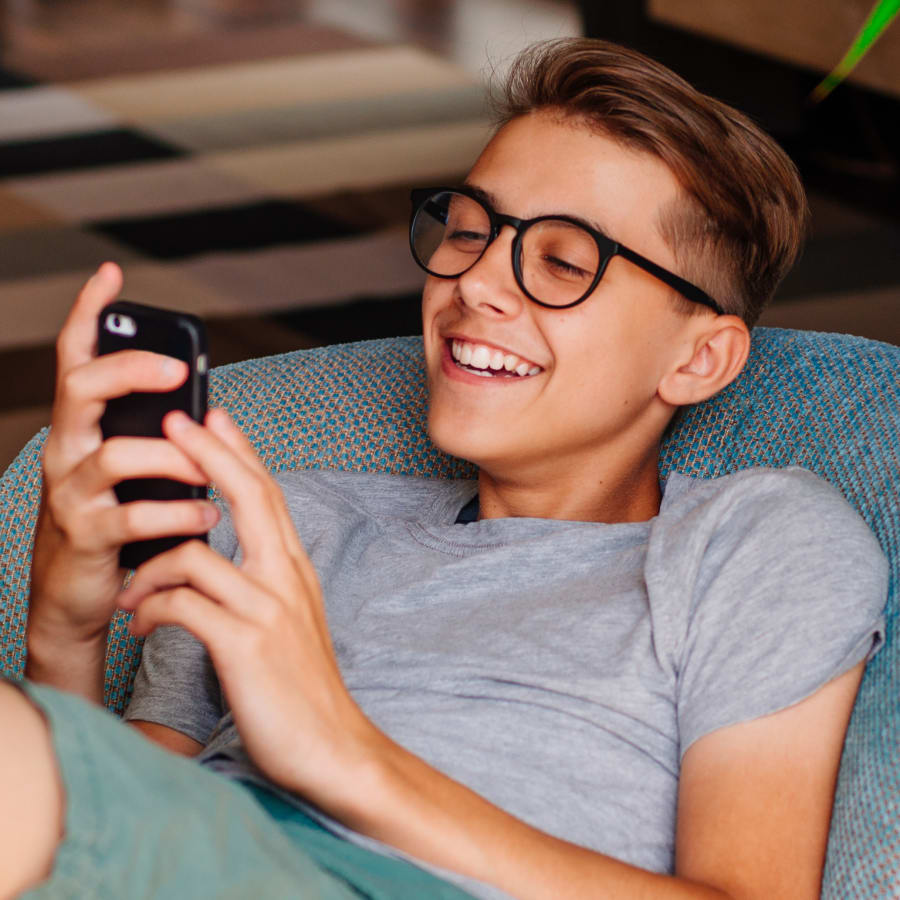 A teenage boy wearing black rimmed glasses, a grey t-shirt, and green shorts sits on a blue bean bag chair smiling while looking at his phone.