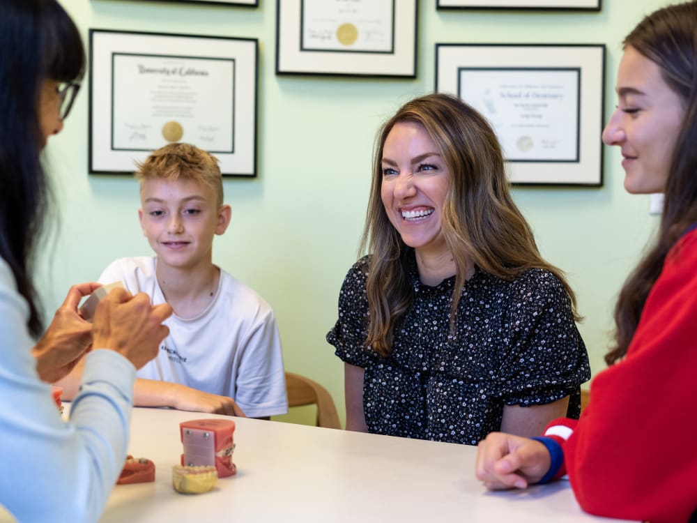 Man, woman and little girl wearing blue and white, sitting on the floor in front of a grey couch laughing together with some plants and bookshelves in the background.